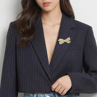 A close-up of a model wearing a dark navy pinstripe blazer with a gold-plated bow-shaped brooch pinned to the chest pocket, paired with light blue jeans.
