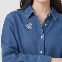 A close-up of a person wearing a blue denim button-up shirt with a cubic zirconia rose brooch on the left chest.