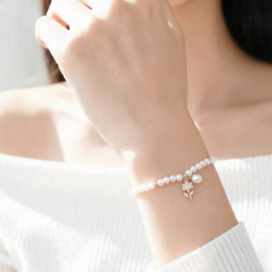 A delicate bracelet worn on a woman's wrist, featuring freshwater pearls and a small rose gold flower charm, shown in a bright, softly lit lifestyle setting.