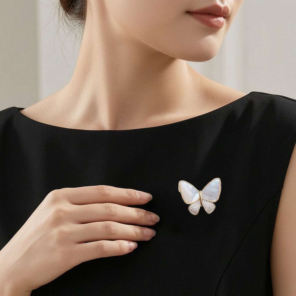 A close-up of a woman wearing a black sleeveless dress , with a butterfly-shaped brooch pinned near the left side of the neckline. The brooch has iridescent mother-of-pearl upper wings with small cubic zirconia on the lower wings.