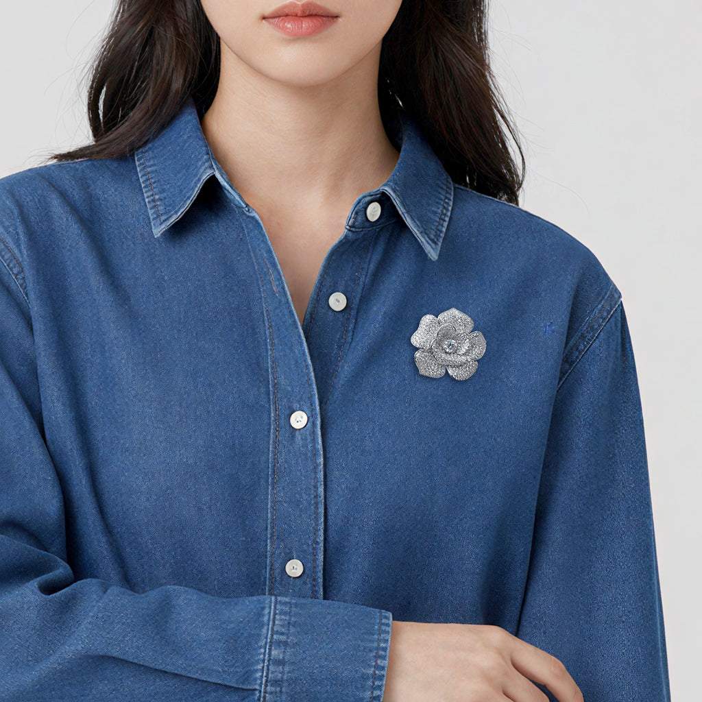 A close-up of a woman wearing a blue denim button-up shirt with a textured silver flower brooch on the chest, set against a plain light gray background.