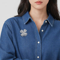 A close-up of a woman wearing a blue denim button-up shirt with a cubic zirconia four-leaf clover brooch pinned to the left chest.
