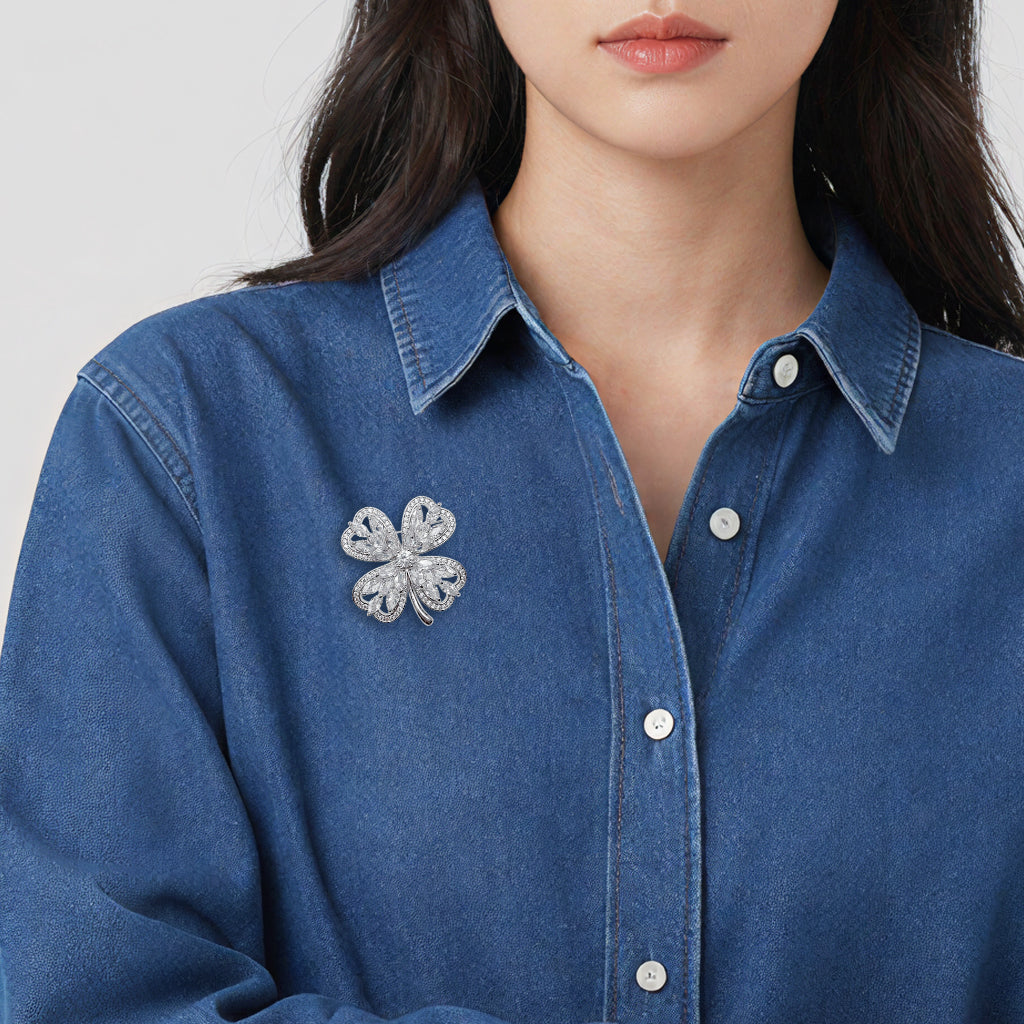 A close-up of a woman wearing a blue denim button-up shirt with a cubic zirconia four-leaf clover brooch pinned to the left chest.