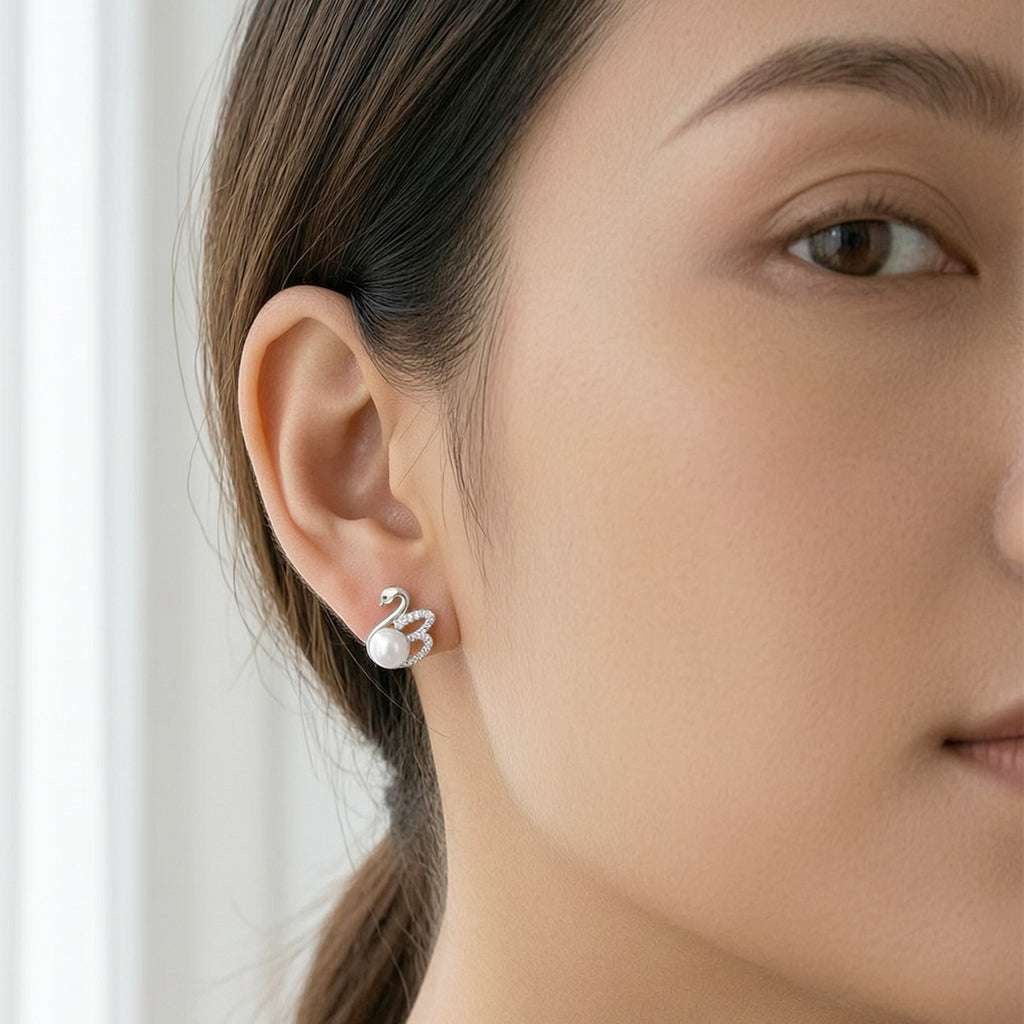 A close-up of a woman's ear wearing a small silver-toned swan stud earring with a freshwater pearl accent.