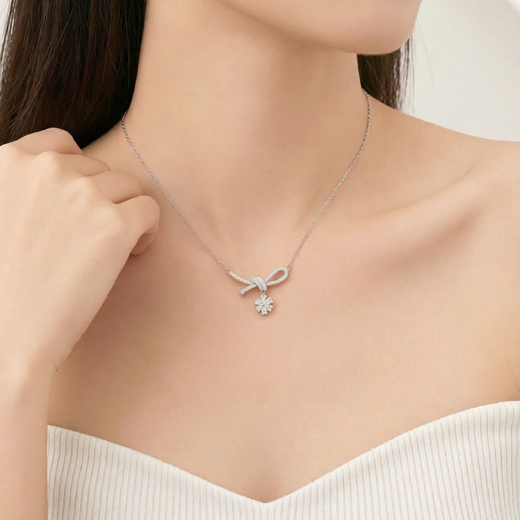 A close-up of a model wearing a delicate silver necklace with a bow and flower pendant, paired with a white ribbed off-the-shoulder top.