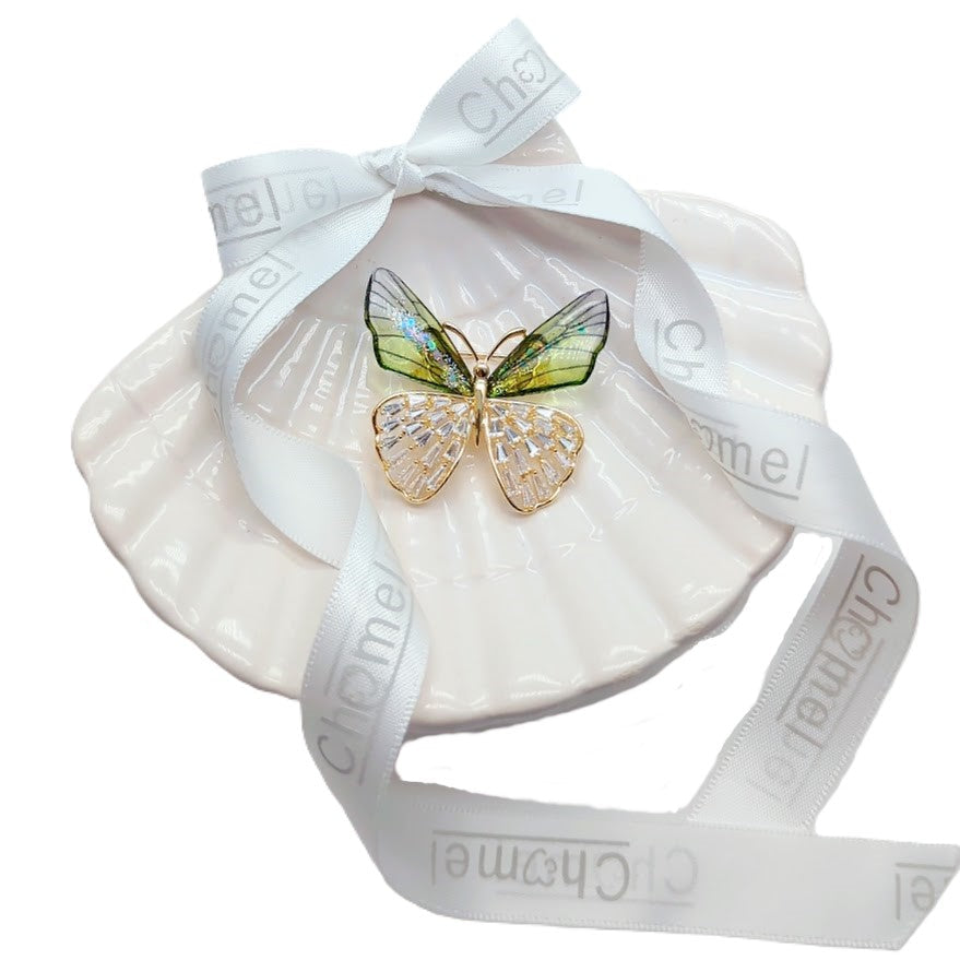 Butterfly cubic zirconia brooch with gold-tone outlines, green iridescent upper wings and clear crystal lower wings, displayed on a ribbon-wrapped shell.