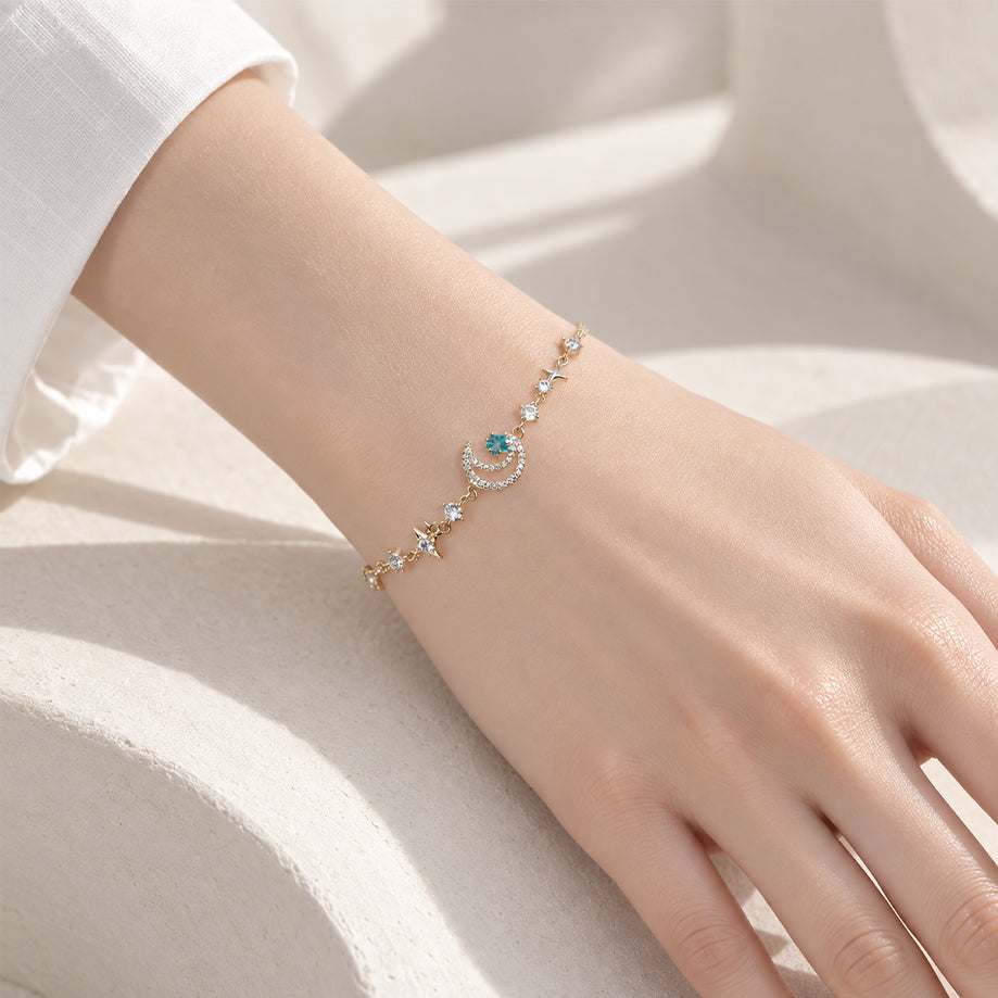 Gold-tone moon bracelet with a delicate chain, clear and turquoise sparkling stones, and small star accents.
